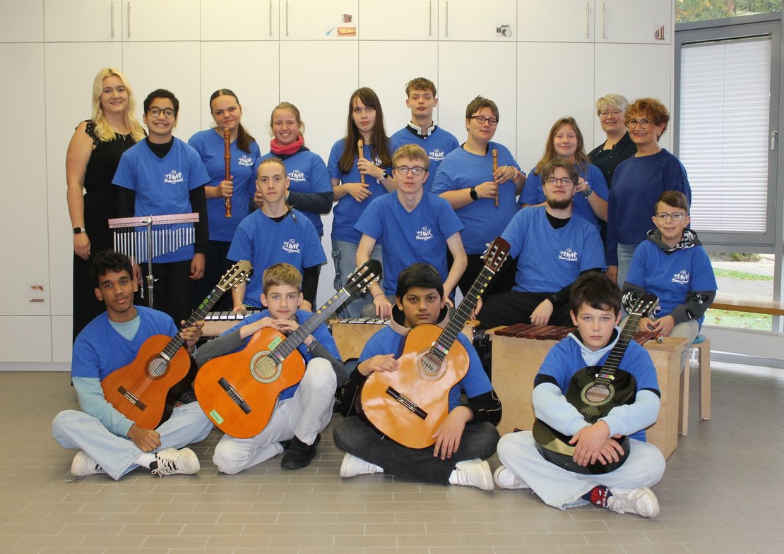 Eine Gruppe von Kindern und Jugendlichen steht und sitzt in mehreren Reihen in einem hellen Raum. Die meisten tragen blaue T‑Shirts mit einem weißen Aufdruck. Vorne sitzen mehrere Kinder auf dem Boden und halten akustische Gitarren in verschiedenen Holzfarben. In der mittleren Reihe sitzen und knien weitere Kinder auf Holzkisten (Cajóns) oder halten Gitarren. In der hinteren Reihe stehen Kinder und Jugendliche mit Blockflöten; links ist außerdem ein Stabspiel zu sehen. Rechts im Hintergrund steht eine große Fensterfront, links und hinten weiße Schränke.