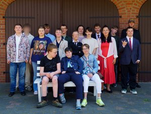 [Leichte Sprache] Gruppenfoto von etwa 17 jungen Erwachsenen vor einem Gebäude mit braunen Holztoren und rotem Backstein. Drei Personen sitzen vorne auf einer weißen Bank, die anderen stehen dahinter in zwei Reihen. Alle tragen festliche Kleidung, zum Beispiel Anzüge, Kleider und Hemden. Eine Person trägt ein T-Shirt mit der Aufschrift 'Abschluss 2026'. Die Gruppe blickt lächelnd in die Kamera.